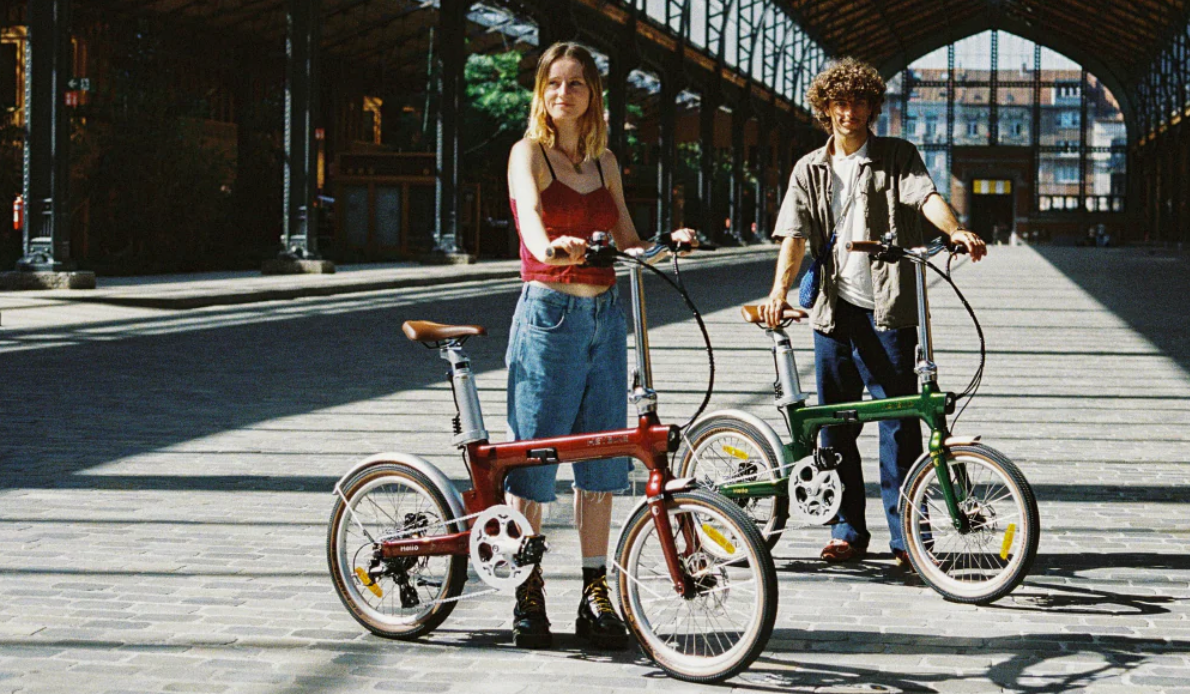 Zwei junge Menschen stehen mit modernen E-Bikes in einer lichtdurchfluteten Bahnhofshalle.