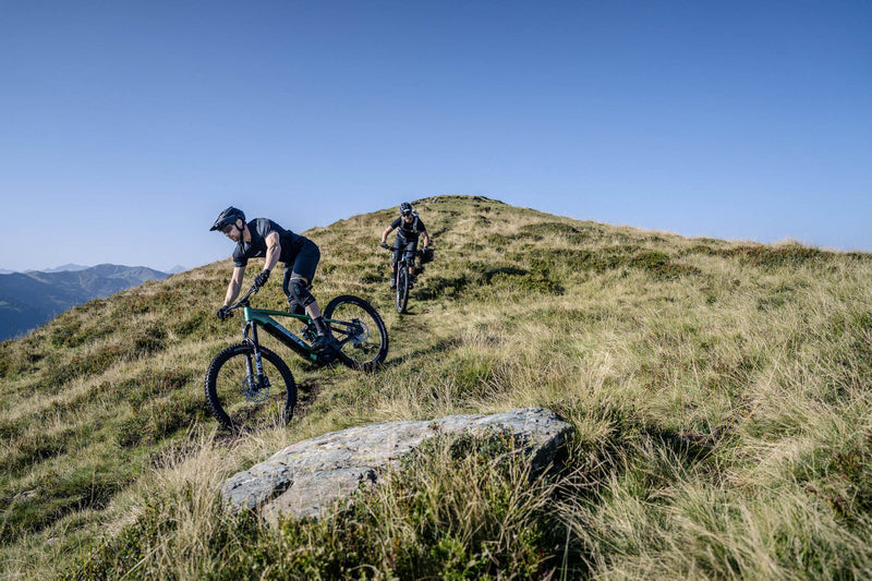 Zwei Mountainbiker fahren mit E-Bikes auf grasigem Berghang bei klarem Himmel.