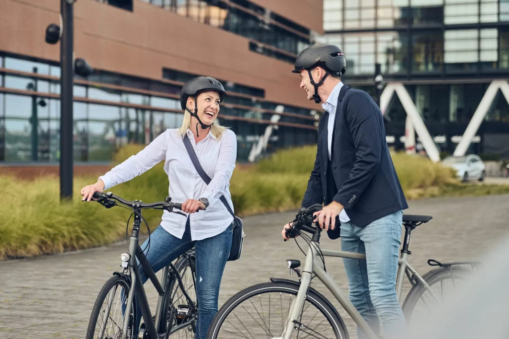 Zwei Erwachsene mit Fahrradhelmen fahren E-Bikes vor modernem Bürogebäude im Freien.